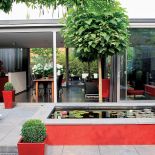 Czerwone akcenty w ogrodzie nawiązują do mebli w kuchni. Zielone kule w wysokich donicach to strzyżony bukszpan. W sadzwce