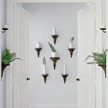 Kompozycja ze starych kinkietów, tyle tylko że zamiast żarówek projektant dodał im białe wazoniki i powkładał w nie liście