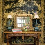 Na ścianie ozdobna tkanina, obok konsola i lustro w złotej ramie.