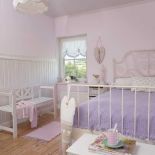 Pastelowe odcienie różu i fioletu, metalowa rama łóżka z dekoracyjnymi wygięciami na szycie, obok ławka