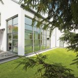 Prosty dom z przeszklonymi ścianami tonie w zieleni.