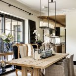 W jadalni rustykalny stół na tle ogromnego okna, a po prawej i lewej czarne kolumny ze stylowymi wazami