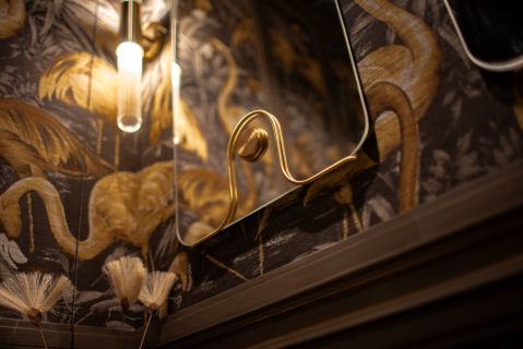 eleganckie wnętrze z czarno-złotą tapetą z motywem flamingów, ozdobnym lustrem w złotej ramię oraz minimalistyczną lampą ścienną o cylindrycznym kloszu