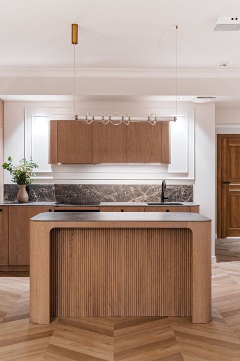 Otwarta kuchnia z zaokrągloną wyspą w stylu warm minimalism. Meble na wymiar wykończone naturalnym fornirem i ciemnym kamieniem.