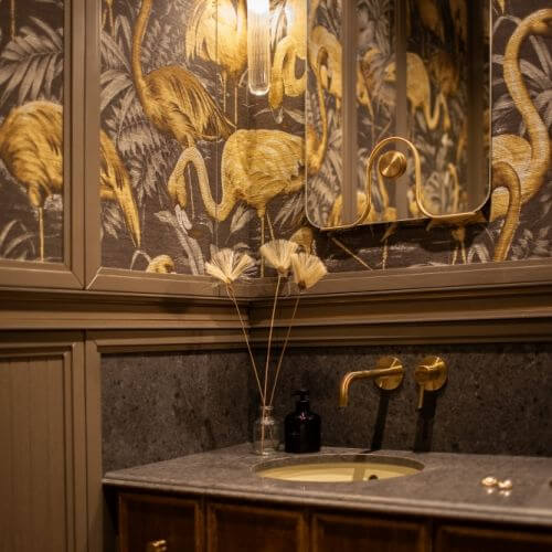 elegancka łazienka z ciemną tapetą w złote flamingi (wzór Flamingo marki Arte), zestawioną z ciemną drewnianą szafką umywalkową oraz złotą armaturą