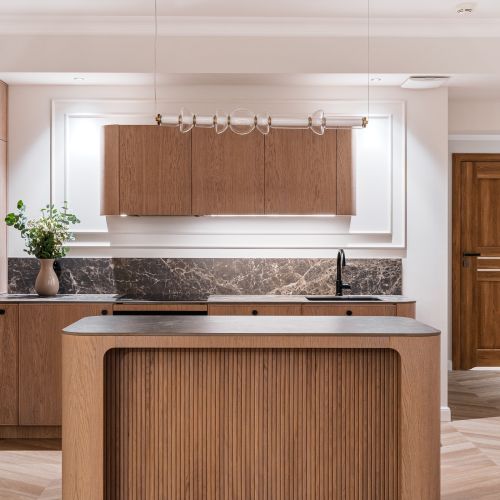 Otwarta kuchnia z zaokrągloną wyspą w stylu warm minimalism. Meble na wymiar wykończone naturalnym fornirem i ciemnym kamieniem.