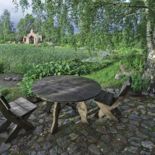 Pod malowniczą brzozą stoją drewniane ławy, krzesła i niski stół.