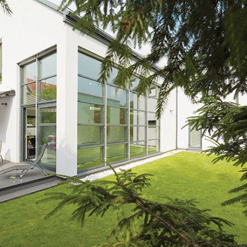 Prosty dom z przeszklonymi ścianami tonie w zieleni.