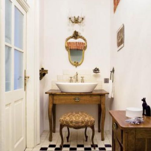 Ściany bielone gruboziarnistym tynkiem, bateria z targu staroci, wiekowa rama lustra, stuletnia kratka do osłonięcia