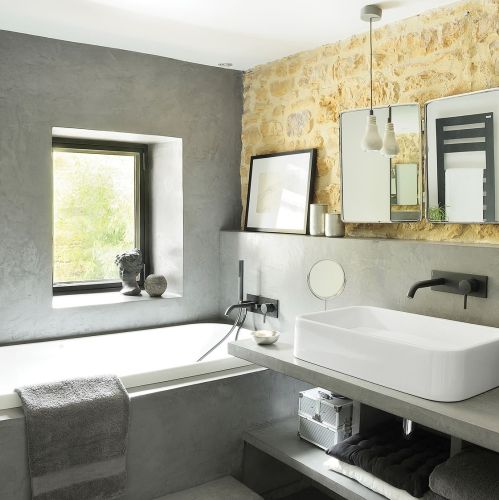 Ściana z nieotynkowanych złocistych kamieni ożywia szarą łazienkę. Minimalizm miesza się z akcentami w stylu