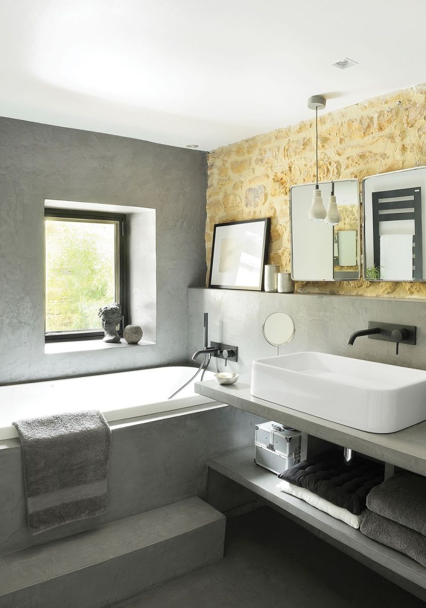 Ściana z nieotynkowanych złocistych kamieni ożywia szarą łazienkę. Minimalizm miesza się z akcentami w stylu