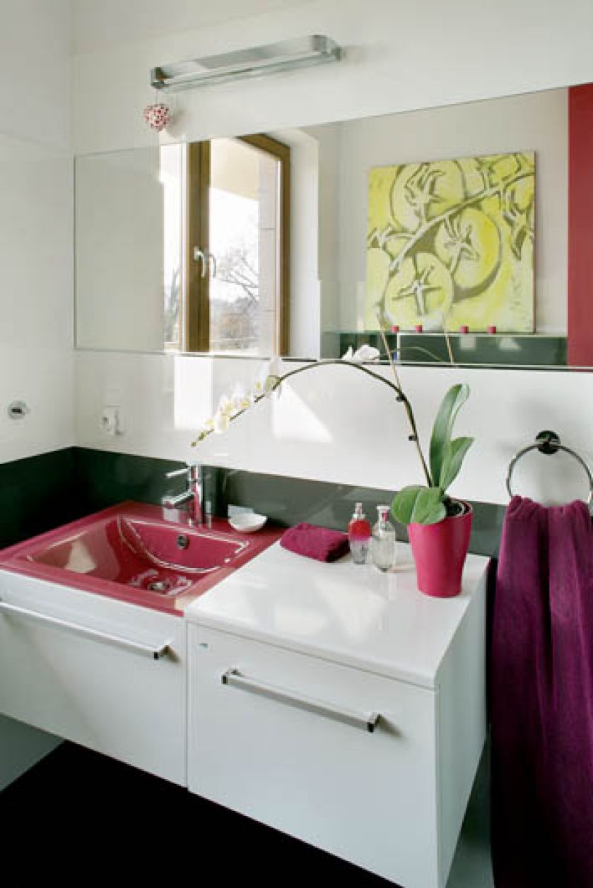 Meble i płytki są biale. Jedyne ekstrawagancje to purpurowa umywalka i kolejny obraz z seriii żółte pomidory.