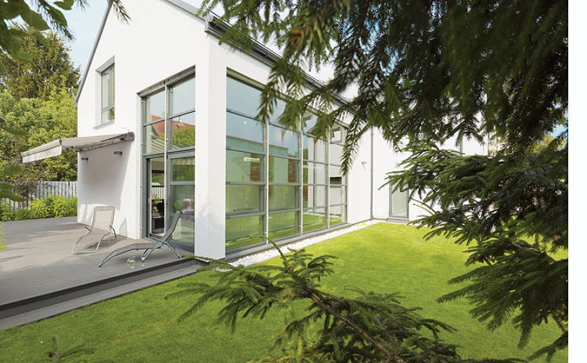 Prosty dom z przeszklonymi ścianami tonie w zieleni.