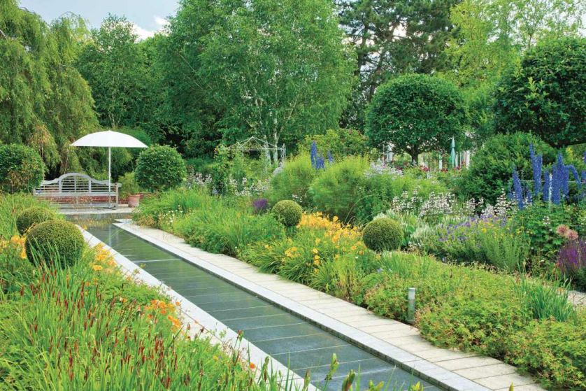 Wokół strumyka rosną symetrycznie posadzone, przycięte starannie krzewy i ogromne kępy kwiatów.