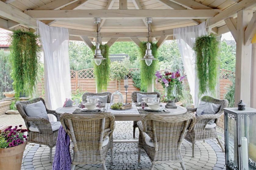 Zadaszony taras w prowansalskim stylu: wiklinowe fotele, duży drewniany stół, brukowa kostka.